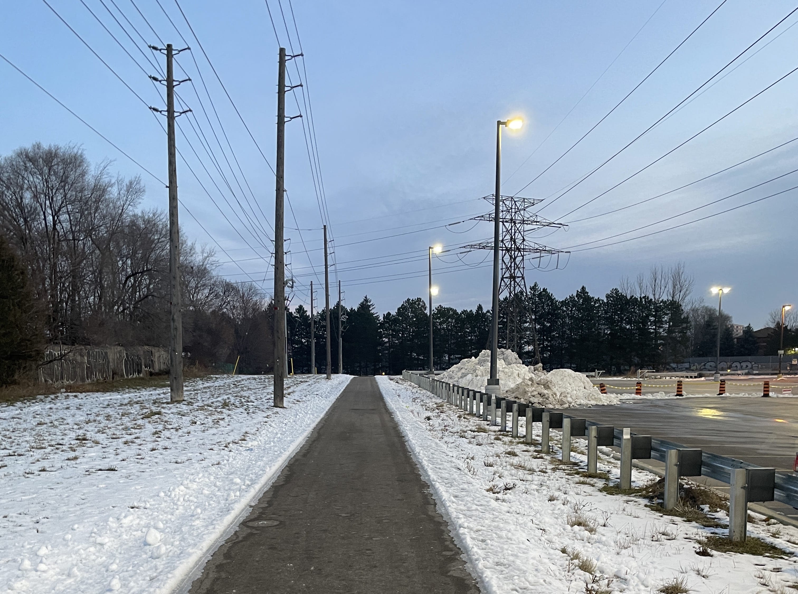 Early-morning photo of the trail near the Scarborough RT line. The black pavement is dry, clear, and visible. Power lines run overhead on both sides.